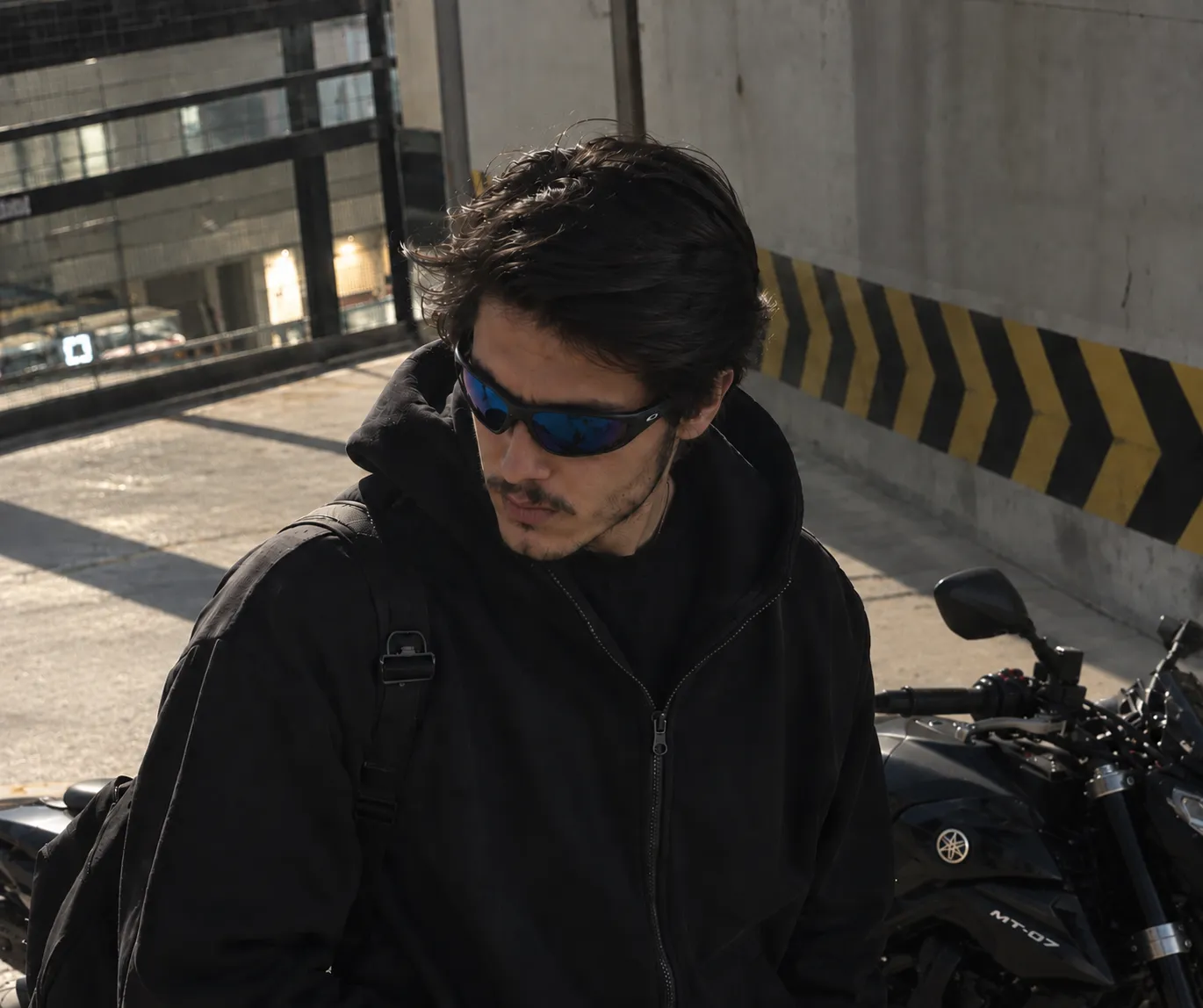 Mustafa; siyah kapüşonlu, güneş gözlüklü, arkasında siyah bir motosiklet.
